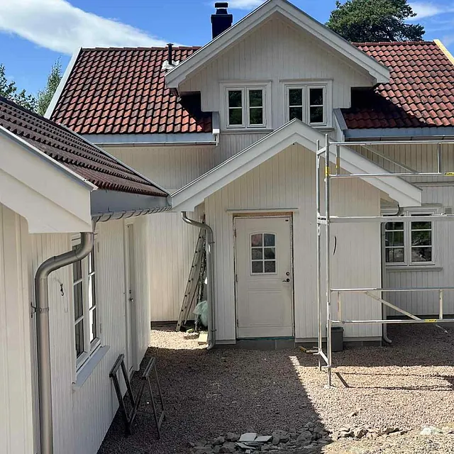 Hvit enebolig med rødt tak og nytt tilbygg, klart for videre arbeid utendørs.