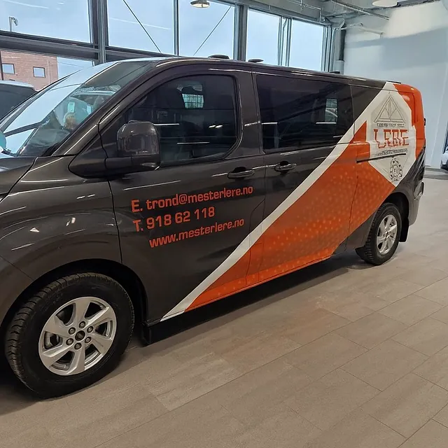 Firmabil med logo og kontaktinfo parkert innendørs, med sort og oransje profilering.