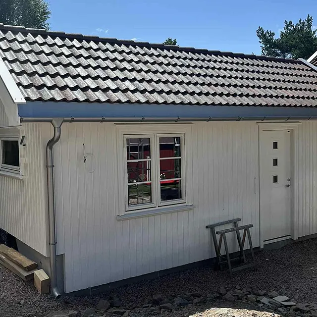 Nybygd tilbygg med grå takstein og hvit panelkledning ved siden av hovedhus.