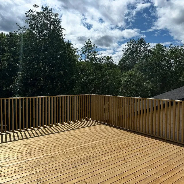 Nybygd terrasse i treverk med rekkverk, omgitt av trær og blå himmel.