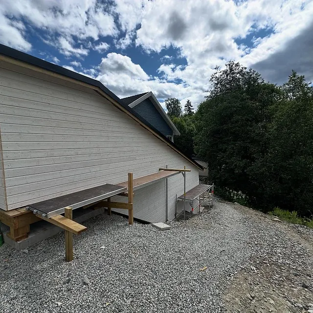 Hvit bolig med tilbygg og gruslagt uteområde, omgitt av trær og blå himmel.