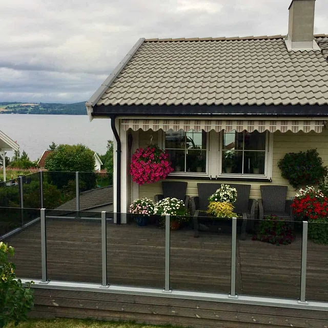 Terrasse med glassrekkverk og blomster foran bolig, med utsikt over vann og landskap.